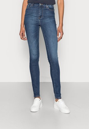Kvinde iført mørkeblå skinny jeans og hvide sneakers, stående mod en ensfarvet lys grå baggrund.