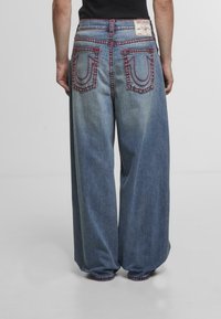 Osoba w luźnych niebieskich dżinsach z czerwonymi szwami i dwiema tylnymi kieszeniami, stojąca na jednolitym jasnym tle.