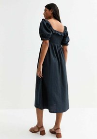 Vestido midi azul oscuro con mangas abullonadas y espalda fruncida. Presenta un escote cuadrado y un corte relajado, combinado con sandalias de tacón marrones.
