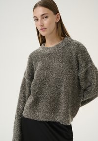 Pullover grigio peloso con una vestibilità ampia, scollatura arrotondata e maniche larghe. Texture morbida con una finitura leggermente lucida. Indossato sopra un indumento nero.