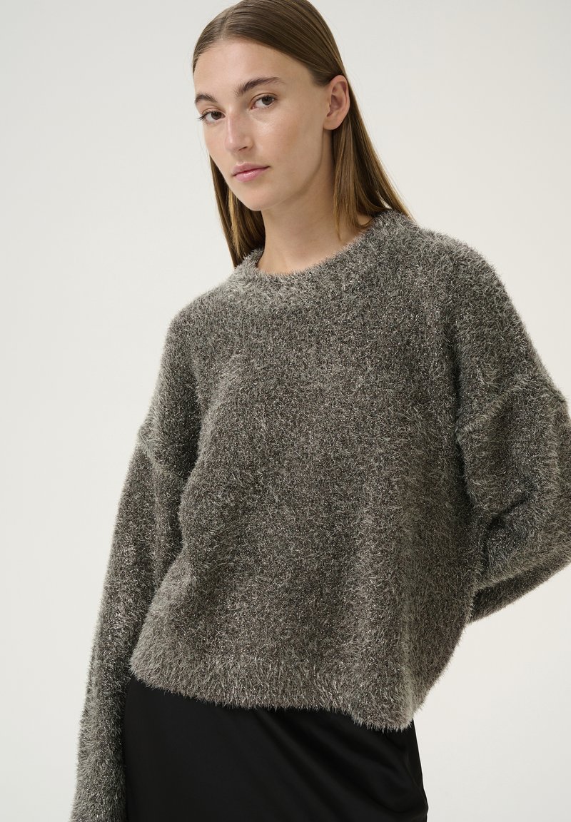 Pullover grigio peloso con una vestibilità ampia, scollatura arrotondata e maniche larghe. Texture morbida con una finitura leggermente lucida. Indossato sopra un indumento nero.