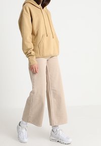 Sudadera beige con bolsillo canguro y cordones, combinada con pantalones de pierna ancha de color claro y zapatillas blancas.