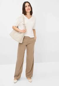 Helle beige übergroße V-Ausschnitt-Bluse, hellbraune weit geschnittene Hosen und eine beigefarbene gewebte Handtasche. Glatter Stoff, lockere Passform und flache cremefarbene Schuhe.