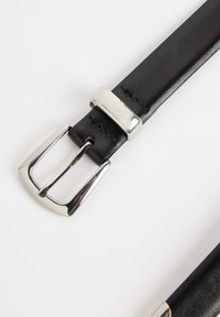 Cinturón de cuero negro con una hebilla de metal plateada brillante. La correa presenta una textura suave con detalles de costura a lo largo de los bordes.