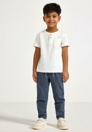 Smilende dreng stående iført en hvid kortærmet skjorte med knapper, blå joggingbukser og hvide sneakers foran en ensfarvet baggrund.