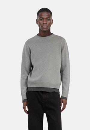 Grauer Strickpullover mit Rundhalsausschnitt und Rippbündchen. Sanfte Textur, lockere Passform, kombiniert mit dunklen Ripp-Hosen.