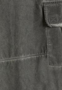 Tessuto grigio scuro con finitura opaca, caratterizzato da cuciture visibili e una tasca applicata. La texture appare leggermente invecchiata.