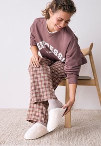 Chaussons confortables en fausse fourrure blanche portés par une personne avec des chaussettes gris clair, assortis à un pantalon de détente à carreaux marron et beige ainsi qu'un sweat-shirt mauve.