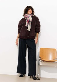 Veste en polaire brun foncé surdimensionnée avec des poignets côtelés, associée à un pantalon large à rayures bleu marine et un foulard à franges multicolores. Talons noirs.