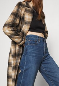 Abrigo de lana a cuadros en tonos beige y negro sobre una camiseta negra corta, combinado con unos vaqueros de mezclilla azul de cintura alta con diseño clásico de cinco bolsillos.