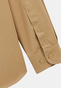 Chemise boutonnée beige avec des poignets pointus, comportant deux boutons marron et une texture de tissu lisse.