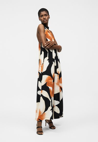 Γυναίκα όρθια με σταυρωμένα τα χέρια φοράει μακρύ μαύρο φόρεμα χωρίς μανίκια με μεγάλα πορτοκαλί και λευκά λουλουδένια σχέδια και μαύρα σανδάλια με τακούνι.