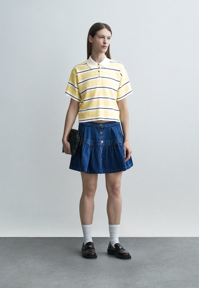 Γυναίκα όρθια που φοράει κοντομάνικη πόλο με κίτρινες και λευκές ρίγες, μπλε τζιν φούστα, λευκές κάλτσες, μαύρα μοκασίνια, κρατώντας μαύρο τσαντάκι χειρός.