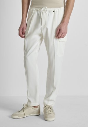 Hombre vistiendo pantalones cargo blancos con bolsillo lateral y zapatillas beige, de pie contra un fondo gris liso.