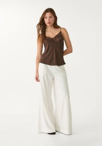 Camisole en satin marron avec bordure en dentelle, associé à un pantalon large blanc. Le mannequin se tient devant un fond clair.