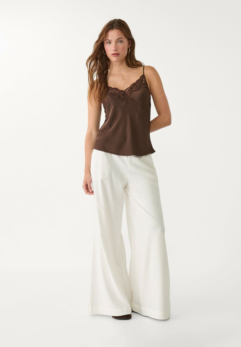 Camisole en satin marron avec bordure en dentelle, associé à un pantalon large blanc. Le mannequin se tient devant un fond clair.