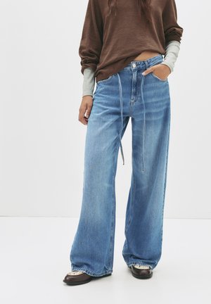 Vaqueros azul claro de pierna ancha con cintura alta, que cuentan con bolsillos delanteros, cremallera y detalle de cordón. Combinados con una prenda superior oversized marrón.