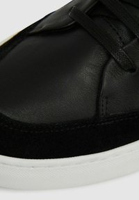 Sneaker nera caratterizzata da una combinazione di pelle liscia e suede testurizzato, con lacci neri e una suola in gomma bianca. Cuciture di dettaglio visibili.