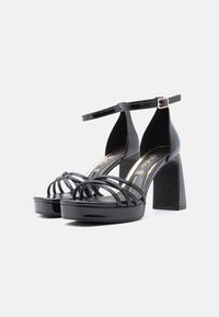 Fekete lakkbőr magas sarkú szandálok pántos kialakítással, nyitott orr-résszel, platform talppal és vastag blokk sarkakkal, bokapántokkal.