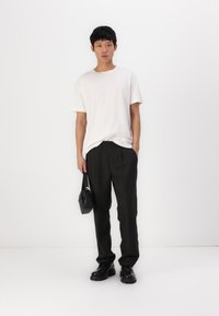 Pantalons noirs avec une taille élastique, associés à un t-shirt blanc ample. Le modèle tient un petit sac noir et porte des chaussures noires imposantes.