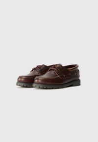 Scarpe da barca in pelle marrone con punta in stile mocassino, cuciture bianche a contrasto e suola in gomma, caratterizzate da un design con lacci e dettagli laterali.