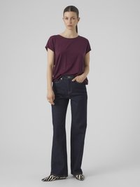 Kvinne som står med mørk lilla kortermet topp, høy midje mørkeblå jeans, zebra-mønstret sko, hår trukket tilbake, nøytral uttrykk.