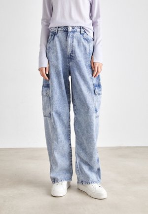Personne portant un jean cargo large bleu clair et des baskets blanches, debout sur un sol clair uni avec un fond blanc.