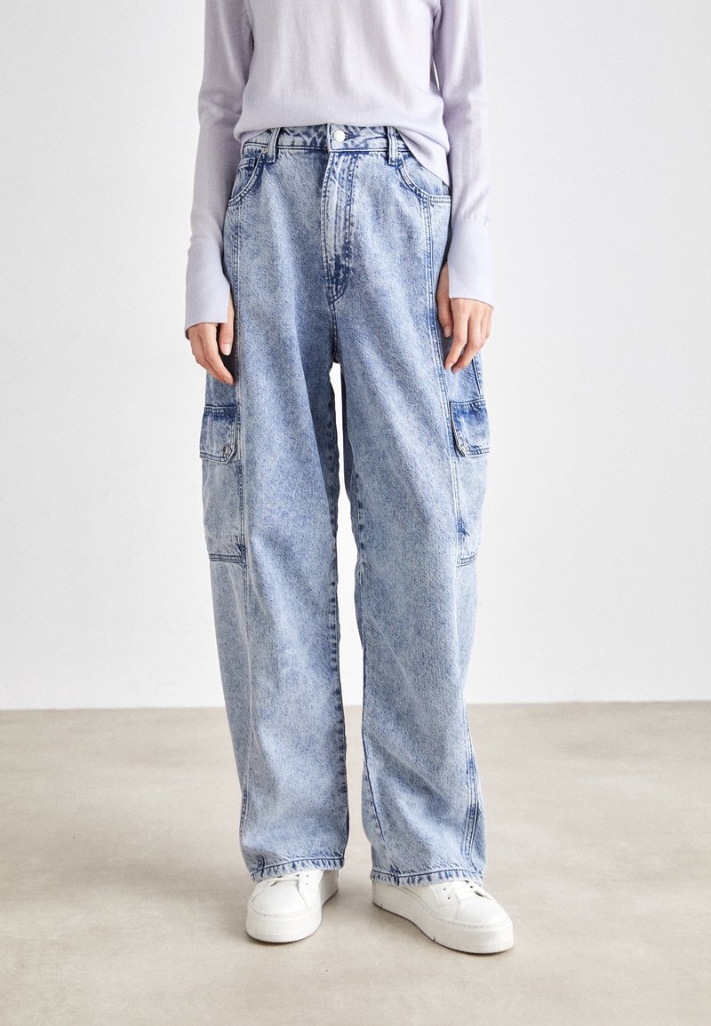 Personne portant un jean cargo large bleu clair et des baskets blanches, debout sur un sol clair uni avec un fond blanc.