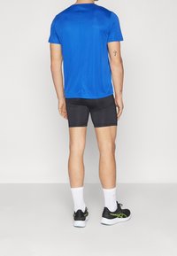 T-shirt de sport bleu, à manches courtes, texture lisse ; associé à un short de compression noir. Chaussures de sport noires et vertes avec des chaussettes blanches.
