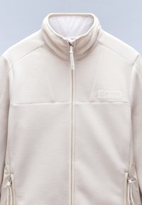 Giacca di pile bianca con colletto alto, zip frontale e tasche laterali. Presenta un tessuto testurizzato e un logo ricamato sul petto.