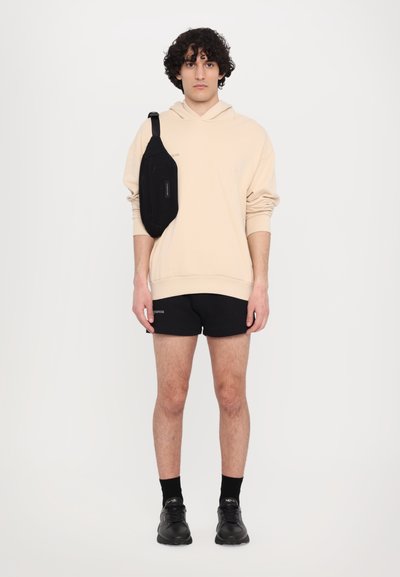 Felpa beige con cappuccio, pantaloni corti neri e sneaker nere. Una piccola borsa a tracolla nera è indossata su una spalla. Design semplice, senza motivi.