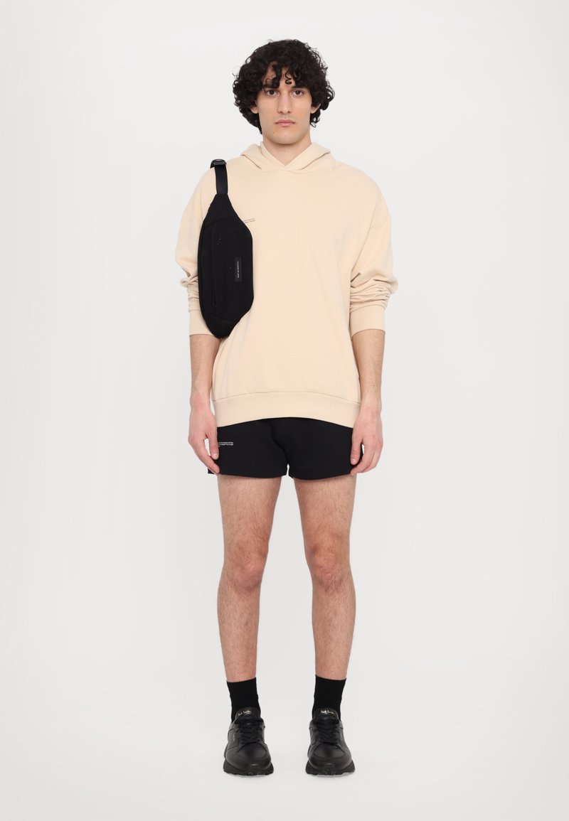 Hanorac bej cu glugă, pantaloni scurți negri și adidași negri. O geantă mică de umăr de culoare neagră este purtată pe un umăr. Design simplu, fără modele.