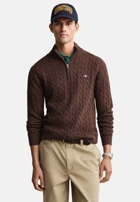 Bruine kabelgebreide trui met een rits bij de hals, gecombineerd met een groene polo en beige broek. Een marineblauwe pet met een geel geborduurd logo.