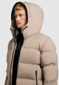 Beige pufferjakke med en stor hætte, har quiltede sektioner, en frontlynlås og to sidelommer med lynlås. Glat, polstret tekstur.