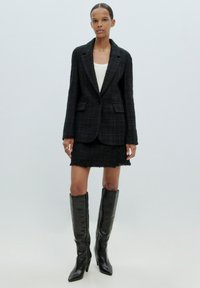 Zwarte tweed blazer met één knoop, gecombineerd met een gestructureerde minirok. Tot de knieën zwarte leren laarzen met een spitse neus en hakken.