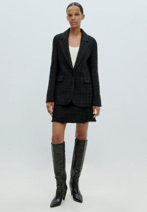 Zwarte tweed blazer met één knoop, gecombineerd met een gestructureerde minirok. Tot de knieën zwarte leren laarzen met een spitse neus en hakken.