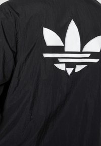 Svart vindjacka med en vit, djärv trebladig logotyp på baksidan, tillverkad av lätt, något strukturerad tyg.