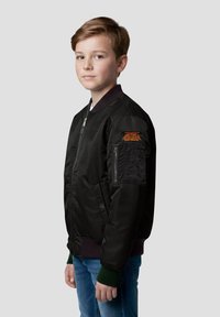 Ragazzo con capelli castani corti che indossa una giacca bomber nera con testo arancione sulla manica e jeans blu, in piedi davanti a uno sfondo grigio semplice.