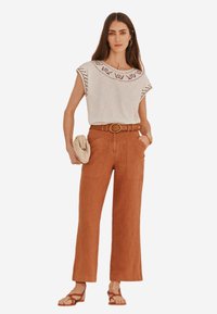 T-shirt gris clair à manches courtes avec un col brodé, associé à un pantalon large couleur rouille, une ceinture marron et des sandales beige.