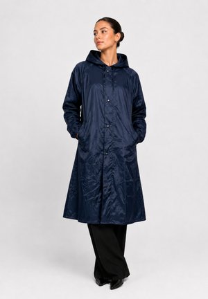 Femme portant un long imperméable bleu marine avec capuche, les mains dans les poches, pantalon noir à jambes larges et chaussures noires pointues, debout devant un fond uni.
