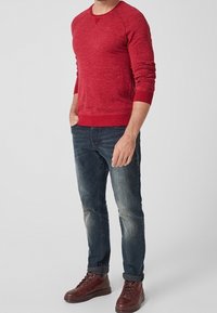 Felpa rossa melange con maniche raglan, polsini a costine e vita a costine. Indossata con jeans blu scuro e stivali marroni con lacci.