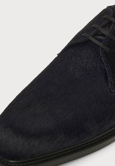 Gros plan d’une chaussure de ville noire avec un matériau texturé ressemblant à des poils et des lacets noirs sur un fond clair.