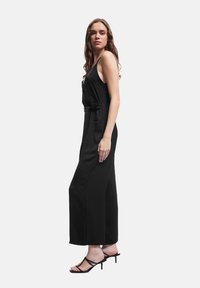 Jumpsuit nero senza maniche con taglio morbido, vita regolabile con cintura, gambe larghe e tasche laterali; la modella indossa tacchi neri a strappo.