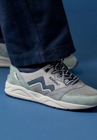 Grå och ljusblå sneaker i mocka och mesh-material; har en vit sula, randiga snören och "Karhu"-logotyp på sidan.