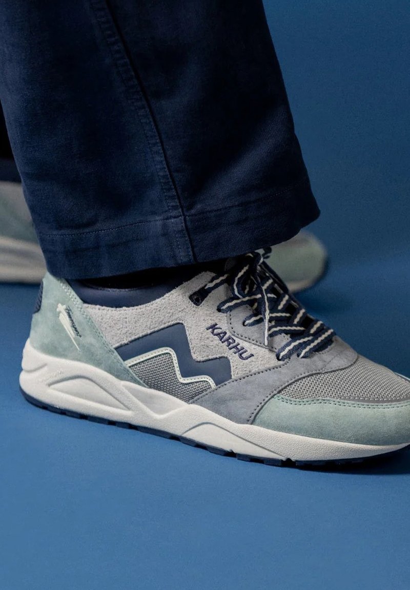 Grå och ljusblå sneaker i mocka och mesh-material; har en vit sula, randiga snören och "Karhu"-logotyp på sidan.