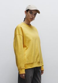 Sudadera amarilla de mangas largas y puños acanalados, combinada con una gorra beige. El modelo se encuentra de pie contra un fondo blanco liso.