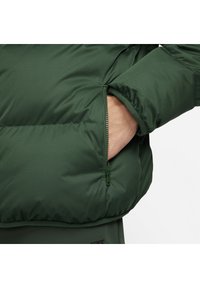 Zielona kurtka puchowa z zamkiem błyskawicznym z lewej strony, o gładkiej, teksturowanej powierzchni i elastycznych mankietach.