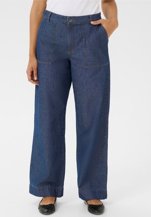 Osoba nosząca niebieskie dżinsy z wysokim stanem i szerokimi nogawkami, z przednimi naszywanymi kieszeniami, w połączeniu z czarnymi balerinami i białym topem.