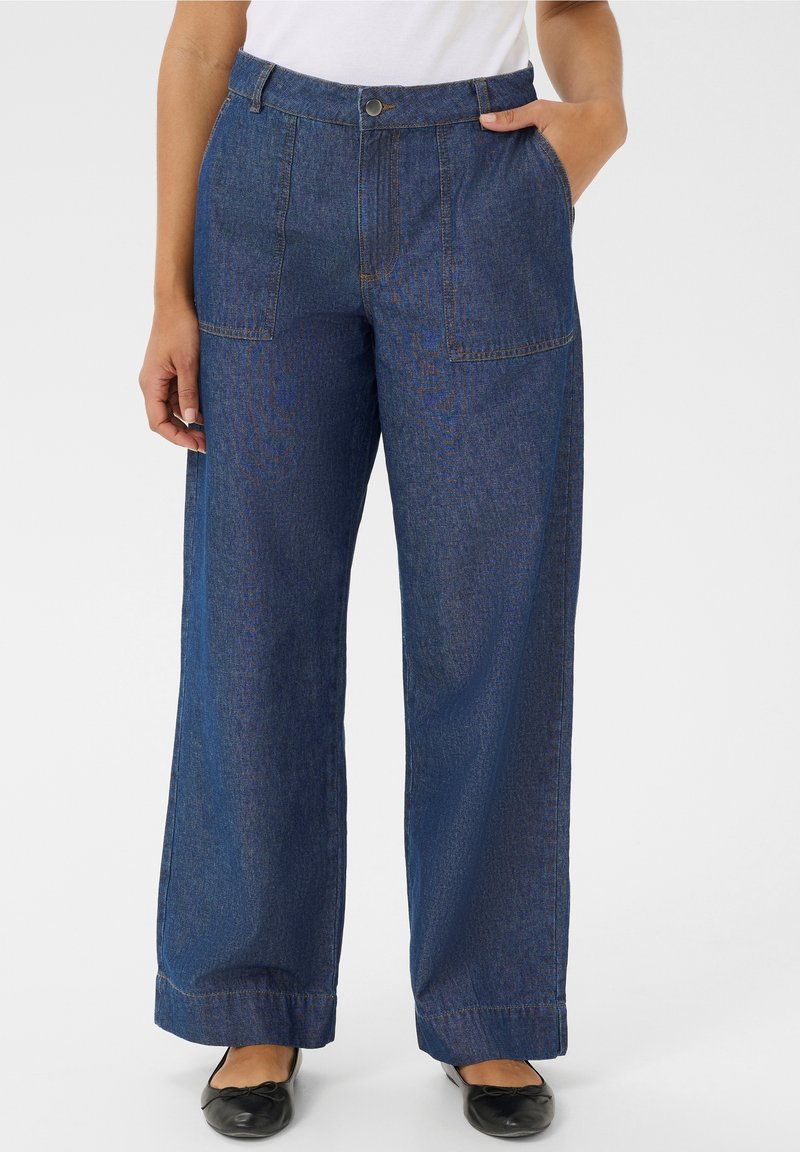 Personne portant un pantalon en jean bleu taille haute à jambes larges avec des poches plaquées sur le devant, associé à des ballerines noires et un haut blanc.