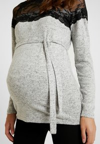 Haut de maternité gris à manches longues, avec un empiècement en dentelle noire, un lien ajustable à la taille et un tissu doux et texturé.